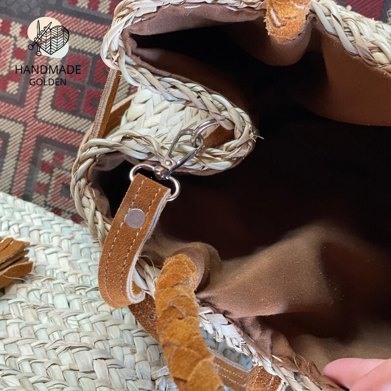 May include: A woven straw handbag with a brown leather strap and braided handle. The interior is lined with brown fabric. The bag features a silver-colored clasp and the text "HANDMADE GOLDEN".