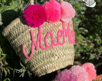 May include: A woven straw tote bag with a handle, adorned with three pink pom-poms and the name "Noela" embroidered in pink yarn. The bag is set against a backdrop of green foliage.