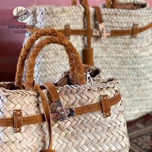 May include: Two woven straw tote bags with brown leather accents. The bags feature braided leather handles, leather straps, and metal clasps. The bags have a natural, neutral color palette, with the text "HANDMADE GOLDEN" in the upper left corner.