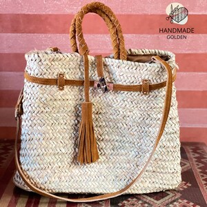 May include: Woven straw tote bag with brown leather accents. Features a braided handle, a shoulder strap, and a decorative tassel. The bag has a rectangular shape and is secured with a leather strap and buckle. The text "HANDMADE GOLDEN" is visible.