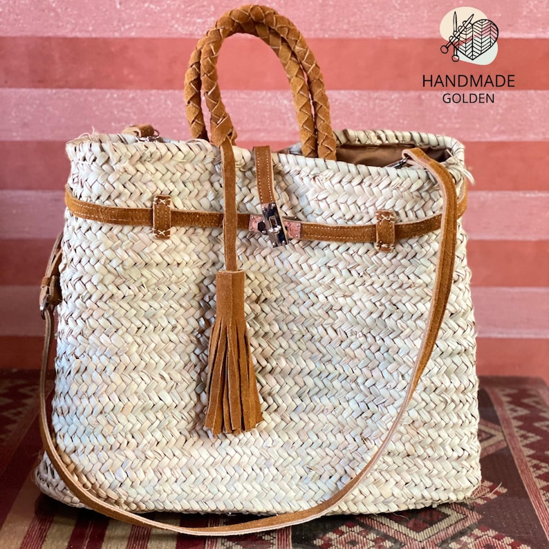 May include: Woven straw tote bag with brown leather accents. Features a braided handle, a shoulder strap, and a decorative tassel. The bag has a rectangular shape and is secured with a leather strap and buckle. The text "HANDMADE GOLDEN" is visible.