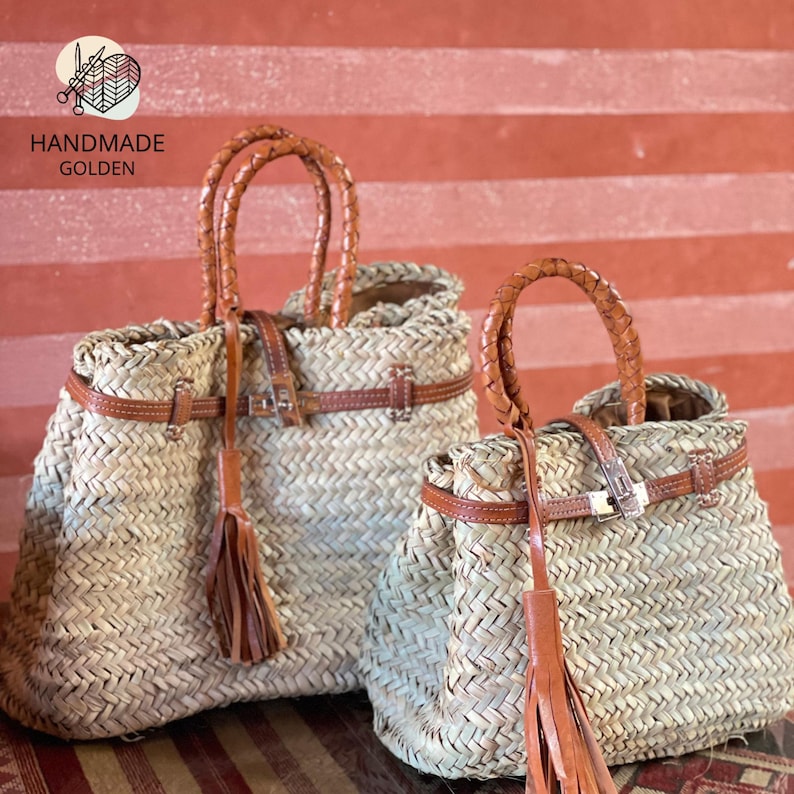 Puede incluir: Dos bolsos de mano de paja tejida con detalles de cuero marr&oacute;n. Los bolsos tienen asas trenzadas, correas de cuero con hebillas y borlas decorativas. Los bolsos son hechos a mano y tienen el texto "HANDMADE GOLDEN".