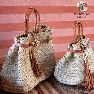 Puede incluir: Dos bolsos de paja trenzada con asas, correas y borlas de cuero marr&oacute;n. Los bolsos tienen un cierre dorado y est&aacute;n etiquetados como "HANDMADE GOLDEN". Los bolsos son de diferentes tama&ntilde;os.