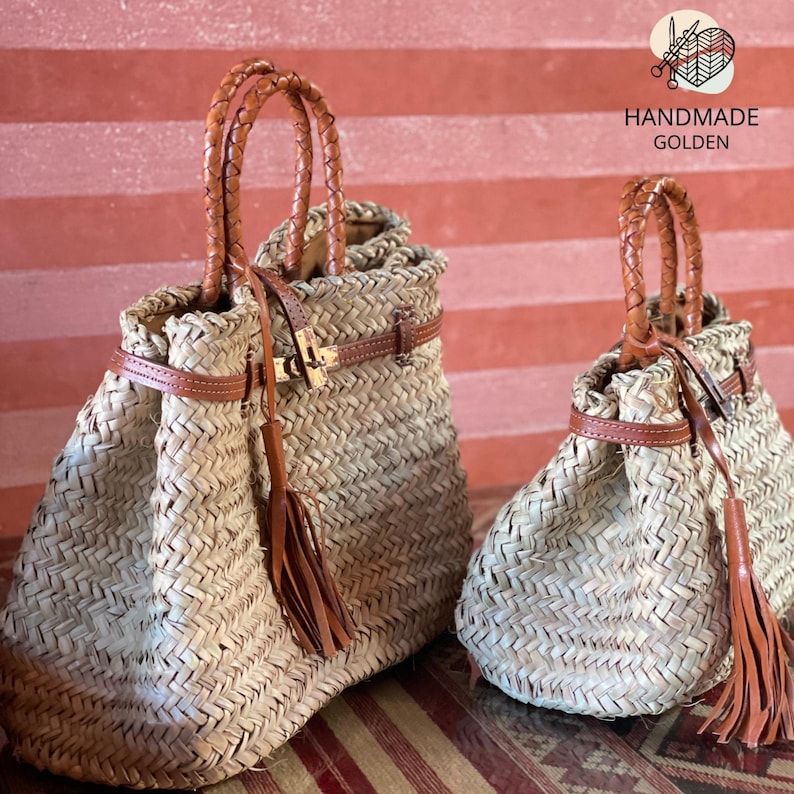 Puede incluir: Dos bolsos de paja trenzada con asas, correas y borlas de cuero marr&oacute;n. Los bolsos tienen un cierre dorado y est&aacute;n etiquetados como "HANDMADE GOLDEN". Los bolsos son de diferentes tama&ntilde;os.