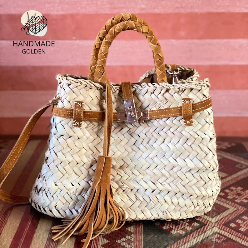 May include: Woven straw handbag with brown leather accents. Features a braided handle, shoulder strap, and a decorative tassel. The bag has a metal clasp closure and leather straps. The text "HANDMADE GOLDEN" is visible in the upper left corner.