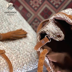 May include: A handmade woven straw tote bag with brown leather straps and accents. The bag features a braided straw trim and a brown interior. The image also shows a matching straw tassel and the text "HANDMADE GOLDEN".