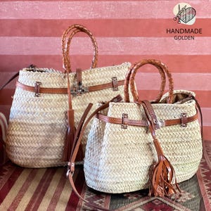 May include: Two woven straw handbags with brown leather accents. The bags have braided handles, leather straps, and decorative tassels. The bags are in different sizes, with the larger one in the background and the smaller one in the foreground. The bags are labeled "HANDMADE GOLDEN".