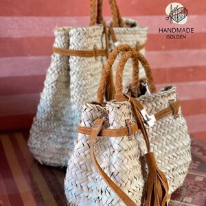 May include: Woven straw handbags with brown leather accents. The bags have braided handles and decorative tassels. The image includes the text "HANDMADE GOLDEN". The bags are in front of a red and brown striped background.
