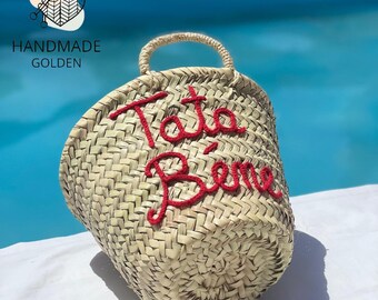 Puede incluir: Un bolso tote de paja tejida con forma redondeada y asa. El bolso presenta texto bordado en rojo que dice "Tata Bien". El bolso es hecho a mano y tiene un color natural.