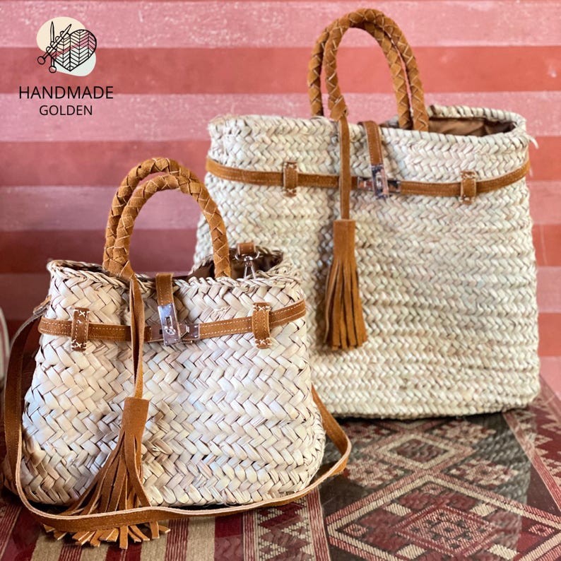 May include: Two woven straw handbags with brown leather accents. The bags have braided handles, leather straps, and decorative tassels. The smaller bag has a shoulder strap. The text "HANDMADE GOLDEN" is visible.