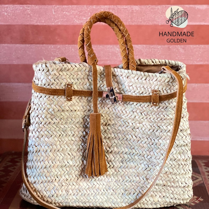 May include: Woven straw tote bag with brown leather accents. Features a braided handle, shoulder strap, and a decorative tassel. The bag has a rectangular shape and is secured with a buckle closure. The text "HANDMADE GOLDEN" is visible on the upper right corner.