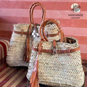 Puede incluir: Dos bolsos de mano de paja tejida con detalles de cuero marr&oacute;n. Los bolsos tienen asas trenzadas, correas de cuero y borlas decorativas. Los bolsos son de color natural, con un cierre met&aacute;lico y el texto "HANDMADE GOLDEN".