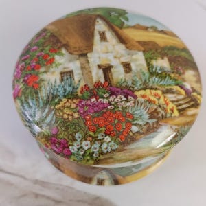 May include: A round, decorative trinket box with a detailed painting of a cottage and garden. The cottage has a thatched roof and is surrounded by colorful flowers in shades of red, pink, and yellow. The box has a glossy finish.