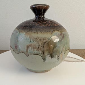May include: A handcrafted ceramic vase with a rounded body and a narrow neck. The vase features a gradient of colors, transitioning from a dark brown top to a light green base, with hints of brown and tan. The vase is a decorative item.