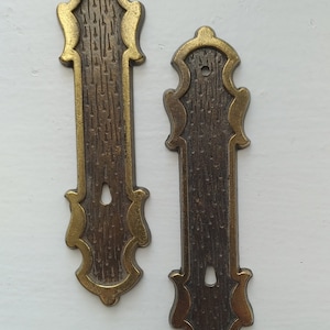 May include: Two ornate brass door or cabinet pulls with a textured finish. The pulls have a gold trim and a keyhole in the center.