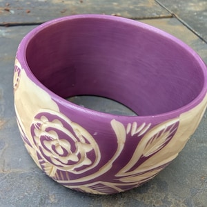 May include: A wide, purple bangle bracelet with carved floral designs. The bracelet features a cream-colored, carved rose and leaf pattern on the outer surface. The interior of the bracelet is a solid, deep purple color. The bracelet is approximately 3 inches wide.