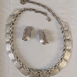 May include: A silver-toned jewelry set consisting of a necklace and a pair of clip-on earrings. The necklace features a series of rectangular, textured metal plates linked by a twisted rope-style chain. The earrings match the rectangular plate design.
