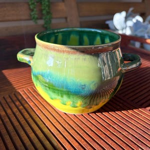 May include: A ceramic bowl with two side handles featuring a glossy glaze in vibrant shades of yellow, teal, and forest green. The colors blend in a drip pattern from the rim down to the base, creating a smooth, reflective surface finish.