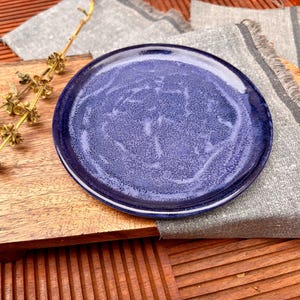 May include: A round, blue ceramic plate with a speckled surface sits on a wooden cutting board. A sprig of dried flowers and a gray linen napkin with fringed edges are also present. The plate has a dark blue rim and a lighter blue center.