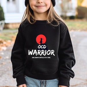 May include: Black sweatshirt featuring a red sun and a silhouette of a person standing on a mountain. The text "OCD Warrior" is printed below the sun, with the phrase "One brave battle at a time." printed below the text.