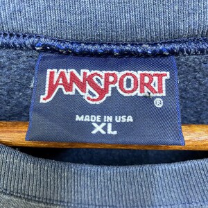 Op de afbeelding: Close-up van een marineblauw sweatshirt met een JanSport-label. Het label is donkerblauw met het JanSport-logo in rood en wit. Op het label staat ook "MADE IN USA" en "XL".