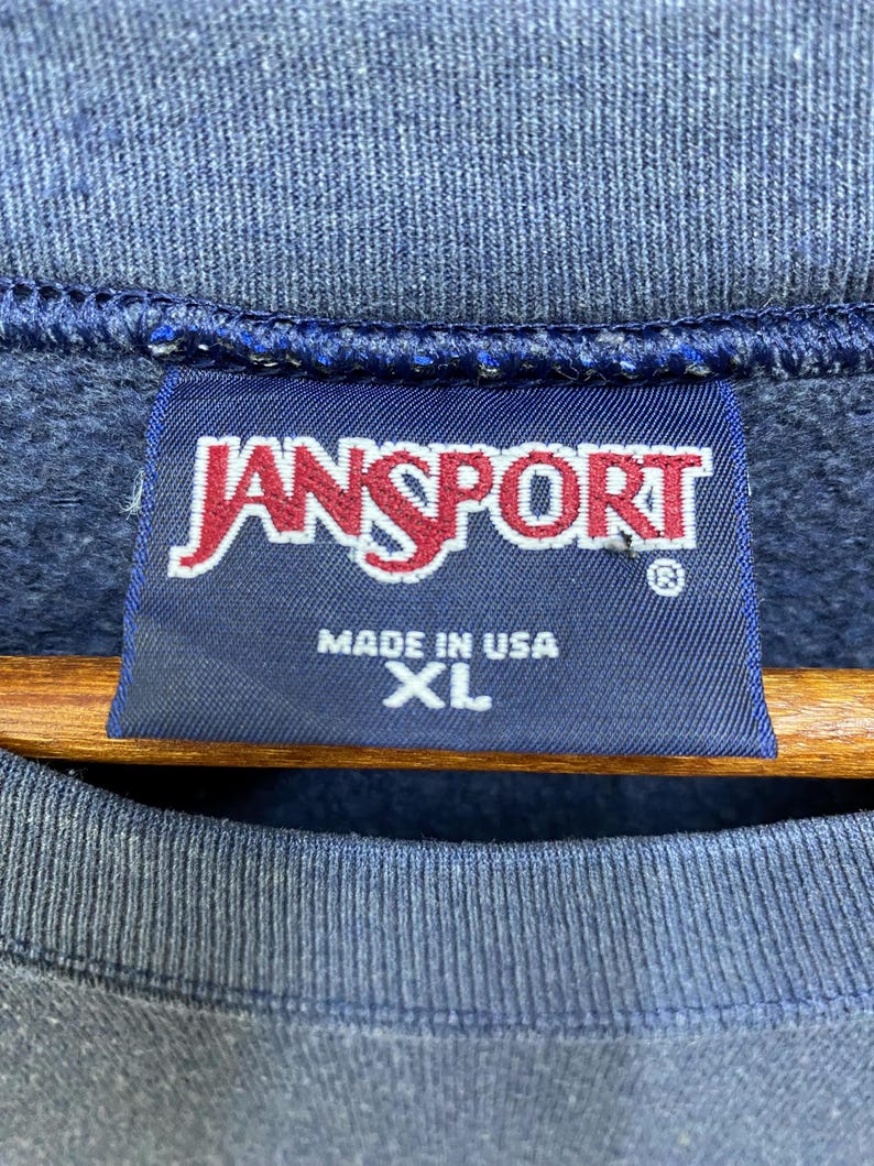 Op de afbeelding: Close-up van een marineblauw sweatshirt met een JanSport-label. Het label is donkerblauw met het JanSport-logo in rood en wit. Op het label staat ook "MADE IN USA" en "XL".