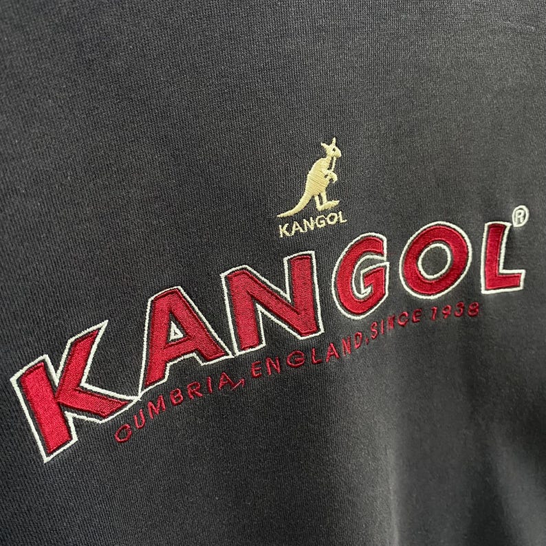 May include: Close-up of a dark grey sweatshirt. The word "KANGOL" is embroidered in red with a white outline. A gold kangaroo logo is above the word "KANGOL". The text "CUMBRIA, ENGLAND, Since 1938" is embroidered below the word "KANGOL".