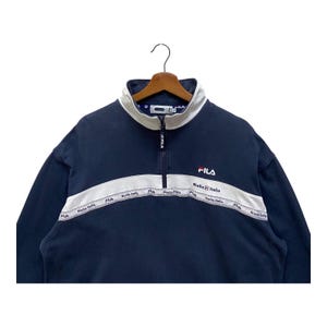 May include: Navy blue FILA sweatshirt with a white collar and a white stripe across the chest. The stripe features the text "Biella Italia". The FILA logo is embroidered in red on the chest.
