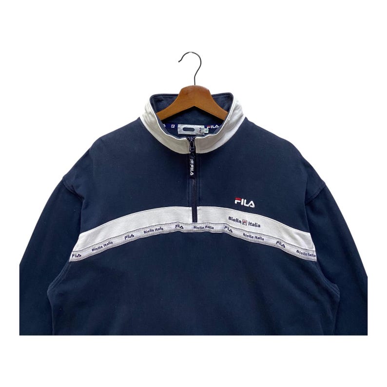 May include: Navy blue FILA sweatshirt with a white collar and a white stripe across the chest. The stripe features the text "Biella Italia". The FILA logo is embroidered in red on the chest.