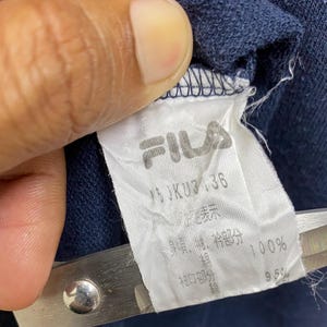 May include: Close-up of a white FILA clothing label with black text and logo. The label is attached to navy blue fabric. The label includes Japanese and English text, with measurements and material percentages.