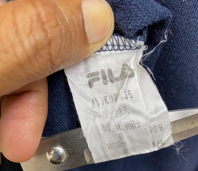May include: Close-up of a white FILA clothing label with black text and logo. The label is attached to navy blue fabric. The label includes Japanese and English text, with measurements and material percentages.