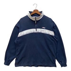 May include: Navy blue long-sleeved sweatshirt with a white collar and a quarter-zip. A white stripe across the chest features the brand name "Fila" and "Italia". The Fila logo is on the right chest.