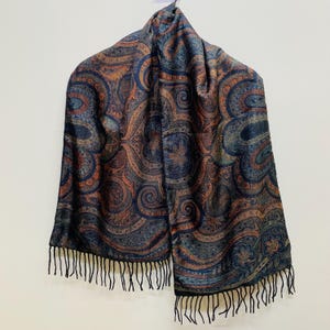 May include: A rectangular scarf featuring a dark blue and copper-toned paisley pattern with intricate swirling motifs. The fabric has a woven texture and is finished with thin, black fringe tassels along the bottom edge of the garment.