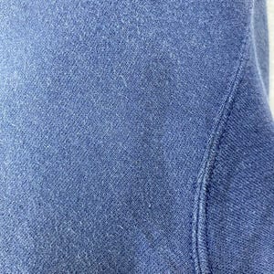 Op de afbeelding: Close-up van een deel van een blauwe sweatshirt. De stof heeft een getextureerde uitstraling met zichtbare vezels. De stiksels langs de naad zijn zichtbaar. De algehele kleur is een gedempt, medium blauw.