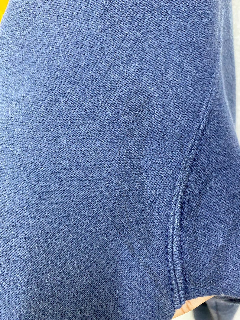 Op de afbeelding: Close-up van een deel van een blauwe sweatshirt. De stof heeft een getextureerde uitstraling met zichtbare vezels. De stiksels langs de naad zijn zichtbaar. De algehele kleur is een gedempt, medium blauw.