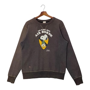 May include: A dark brown sweatshirt with long sleeves, featuring a Snoopy graphic in the center. The graphic includes the text "1st CAV. DIV. AIR BORNE" and a yellow and black shield design. The sweatshirt is hanging on a wooden hanger.