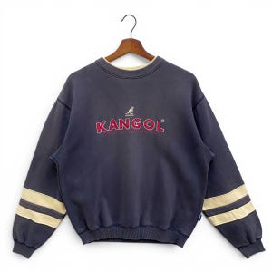 May include: Navy blue Kangol sweatshirt with a red logo and kangaroo graphic. The long sleeves feature two beige stripes. The crew neck has a cream-coloured trim. The sweatshirt is hanging on a wooden hanger.