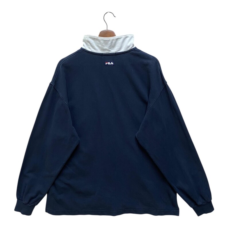 May include: Navy blue long-sleeved shirt with a white collar and the Fila logo. The shirt has a mock neck and is hanging on a wooden hanger. The Fila logo is in red and white.