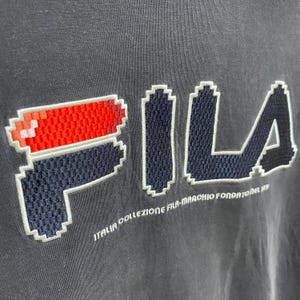 May include: Close-up of a dark grey t-shirt with the brand name "FILA" in a pixelated design. The logo includes red and white accents. Below the logo, the text "ITALIA COLLEZIONE FILA-MARCHIO FONDATOREL 1911" is visible.