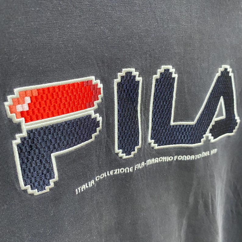 May include: Close-up of a dark grey t-shirt with the brand name "FILA" in a pixelated design. The logo includes red and white accents. Below the logo, the text "ITALIA COLLEZIONE FILA-MARCHIO FONDATOREL 1911" is visible.