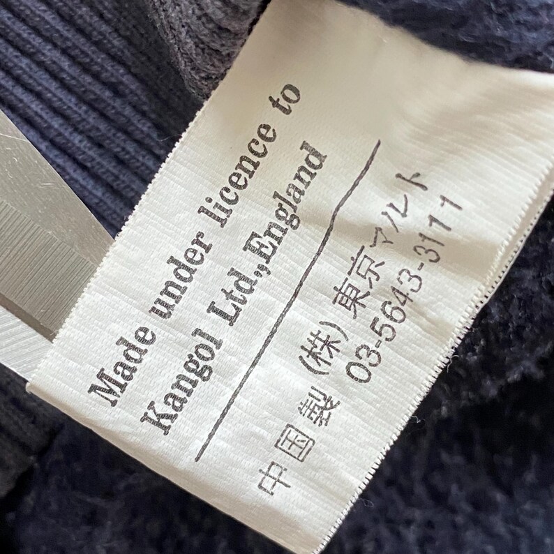 May include: A white label with black text, partially visible, reads "Made under licence to Kangol Ltd., England." Japanese text and numbers are printed below. The label is against a dark blue fabric.
