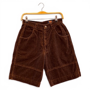 May include: Brown corduroy shorts hanging on a wooden hanger. The shorts have a high waist, front pockets, and a button closure. The fabric has a ribbed texture. The shorts are a medium brown color.