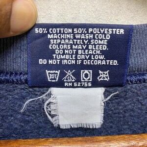 Op de afbeelding: Close-up van een marineblauw kledinglabel met witte tekst. Het label luidt: "50% Katoen 50% Polyester, Machinewas koud, apart. Sommige kleuren kunnen uitlopen. Niet bleken. Drogen in de droger op lage temperatuur. Niet strijken indien versierd." Met wassymbolen en "RN 52755".