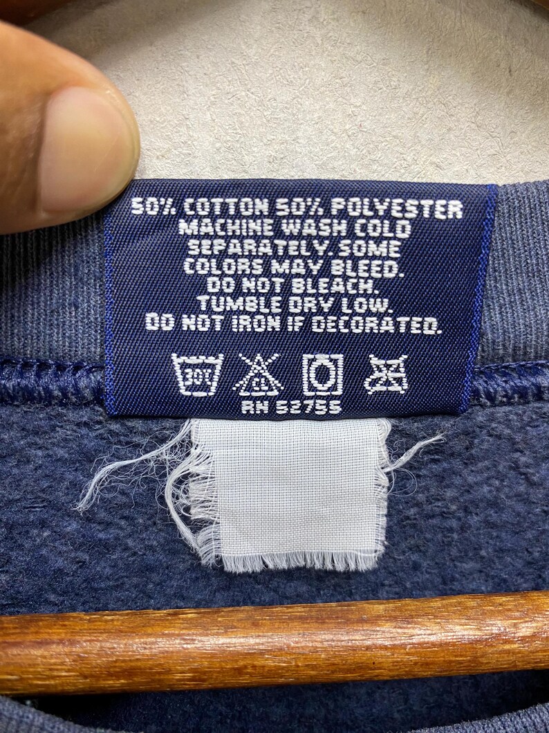 Op de afbeelding: Close-up van een marineblauw kledinglabel met witte tekst. Het label luidt: "50% Katoen 50% Polyester, Machinewas koud, apart. Sommige kleuren kunnen uitlopen. Niet bleken. Drogen in de droger op lage temperatuur. Niet strijken indien versierd." Met wassymbolen en "RN 52755".