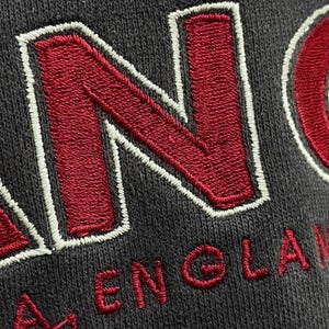 May include: Close-up of a dark grey fabric with embroidered red letters spelling "ENGLAND". The letters are outlined in white thread. The fabric has a textured appearance.