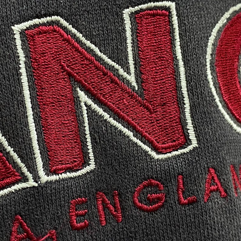 May include: Close-up of a dark grey fabric with embroidered red letters spelling "ENGLAND". The letters are outlined in white thread. The fabric has a textured appearance.