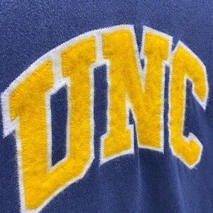 Op de afbeelding: Close-up van een marineblauw sweatshirt met de letters "UNC" in gele viltletters met een witte omlijning. De letters zijn gebogen over de voorkant, wat een collegiale uitstraling geeft.