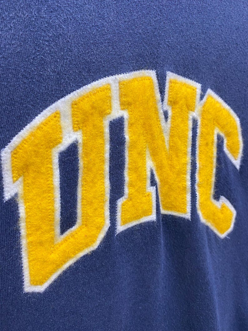 Op de afbeelding: Close-up van een marineblauw sweatshirt met de letters "UNC" in gele viltletters met een witte omlijning. De letters zijn gebogen over de voorkant, wat een collegiale uitstraling geeft.