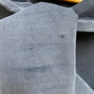 May include: Close-up of a dark grey fabric with visible stains and a small hole. The texture of the material is evident, showing the weave of the fibres. The image highlights the imperfections of the textile.