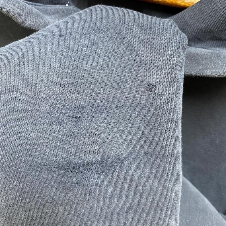 May include: Close-up of a dark grey fabric with visible stains and a small hole. The texture of the material is evident, showing the weave of the fibres. The image highlights the imperfections of the textile.